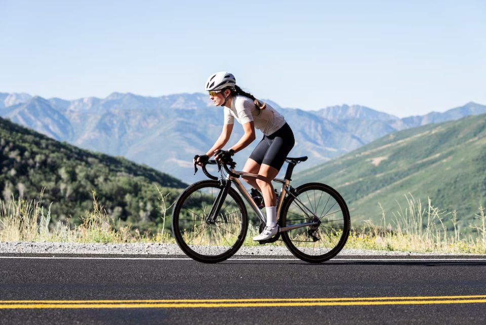 woman in black shorts and a white t-shirt riding a bike on the road, hills, and mountains behind